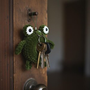 May include: A green crocheted frog keyring with large white and black eyes hangs on a wooden door. The frog holds a set of keys on a metal ring. The door has a brass doorknob and a dark brown finish.