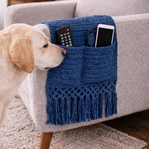 Può includere: Un organizer per divano all'uncinetto blu navy con tasche e frange. Un telecomando, occhiali e uno smartphone sono nelle tasche. Un labrador giallo è accanto al divano.