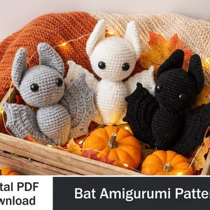 Pode incluir: Três amigurumis de morcego de crochê em uma caixa de madeira com abóboras e folhas de outono. Os morcegos são cinza, branco e preto. O texto "Digital PDF Download" e "Bat Amigurumi Pattern" é visível.