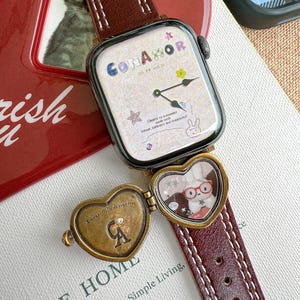 Peut inclure: Une montre connectée carrée avec un bracelet en cuir bordeaux et des coutures blanches. Le cadran affiche le mot "CONAMOR" et un petit motif floral. Un médaillon en forme de cœur avec un lapin est attaché.