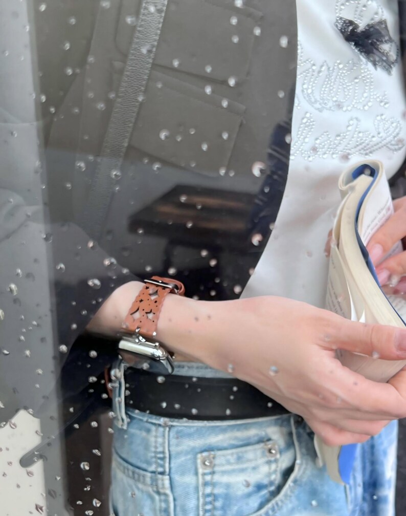 May include: A brown leather watch band with a cut-out design is attached to a silver smartwatch. The watch is worn on a wrist, paired with a black jacket, a white top, and blue jeans. The watch band has a buckle closure.