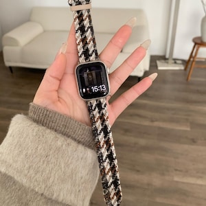 May include: A silver smartwatch with a houndstooth patterned strap in black, brown, and white. The watch face shows the time as 15:13. The strap is attached to a beige leather section.