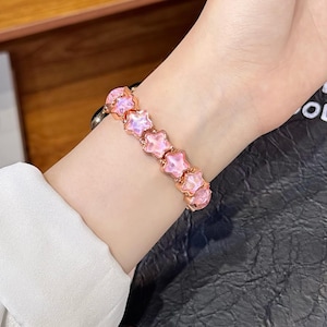 May include: A bracelet featuring a series of connected star-shaped charms with a metallic rose gold finish and translucent, light pink, iridescent centers. The star charms are linked together in a continuous row to form a flexible wrist band.