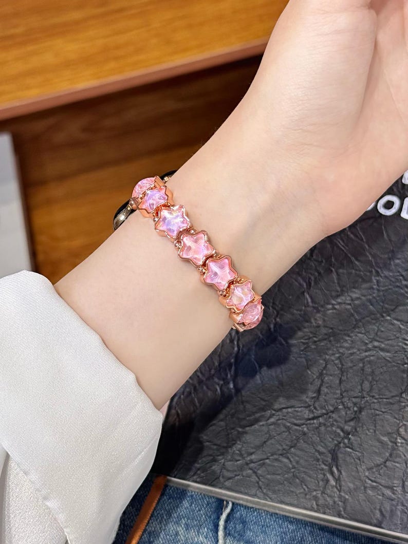 May include: A bracelet featuring a series of connected star-shaped charms with a metallic rose gold finish and translucent, light pink, iridescent centers. The star charms are linked together in a continuous row to form a flexible wrist band.