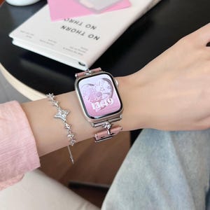 May include: A silver smartwatch with a pink band and a pink heart-shaped display. A silver bracelet with star-shaped charms is also visible. The watch displays the time 13:49. A pink notebook and a white book with the text "NO THORN" are in the background.