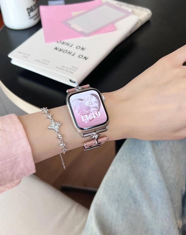May include: A silver smartwatch with a pink band and a pink heart-shaped display. A silver bracelet with star-shaped charms is also visible. The watch displays the time 13:49. A pink notebook and a white book with the text "NO THORN" are in the background.