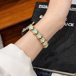 May include: A gold-toned watch band featuring a series of connected star-shaped links with pale mint green centers. The band is attached to a black electronic watch face, creating a decorative accessory that wraps around the wrist.