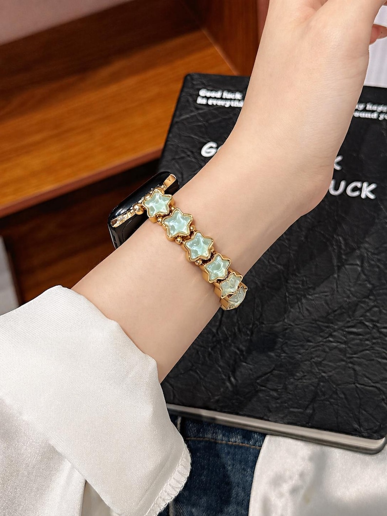 May include: A gold-toned watch band featuring a series of connected star-shaped links with pale mint green centers. The band is attached to a black electronic watch face, creating a decorative accessory that wraps around the wrist.
