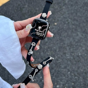 Może przedstawiać: Czarny skórzany pasek do zegarka ze srebrnymi akcentami, w tym ćwiekami w kształcie róży i wiszącymi zawieszkami. Tarcza zegarka wyświetla tekst "Chrome Hearts" i godzinę 19:07. Pasek posiada srebrną klamrę.