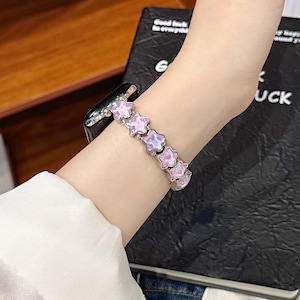 May include: A smartwatch band featuring a series of linked, five-pointed star-shaped charms. Each star has a translucent, iridescent light pink and purple finish with a metallic silver-toned border, connected by small silver-toned metal beads.