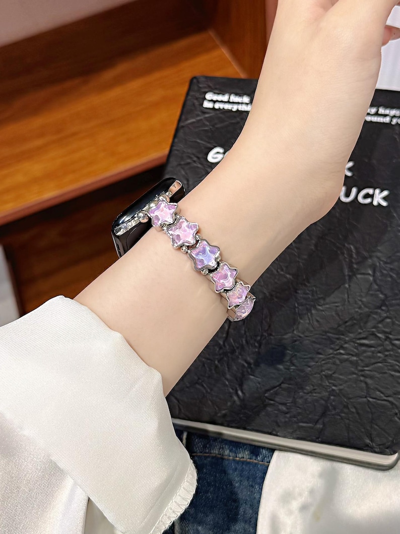 May include: A smartwatch band featuring a series of linked, five-pointed star-shaped charms. Each star has a translucent, iridescent light pink and purple finish with a metallic silver-toned border, connected by small silver-toned metal beads.