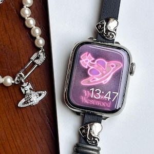 以下が含まれることがあります： 黒い革のバンドと装飾的な金属アクセントが付いたシルバーのスマートウォッチ。時計の文字盤にはピンクのグラフィックと13:47の時刻が表示され、「Westwood」の文字があります。装飾的なチャームが付いたパールネックレスも表示されています。