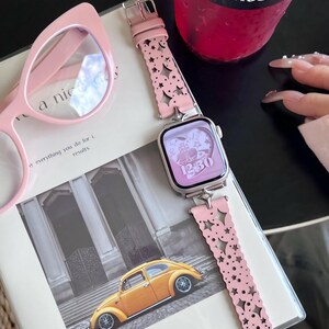 May include: A pink smartwatch with a matching pink band featuring star and moon cutouts. The watch face displays the time as 12:30. Beside the watch are pink cat-eye glasses and a book with a photo of a yellow car. A pink drink is in the background.