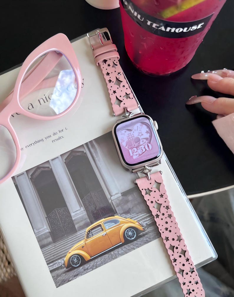 May include: A pink smartwatch with a matching pink band featuring star and moon cutouts. The watch face displays the time as 12:30. Beside the watch are pink cat-eye glasses and a book with a photo of a yellow car. A pink drink is in the background.
