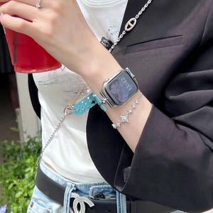 May include: A silver smartwatch with a digital display is worn on the wrist, paired with a star-shaped bracelet. A turquoise watch band and a silver chain are also visible. The person is wearing a black blazer and a black belt with a silver buckle.