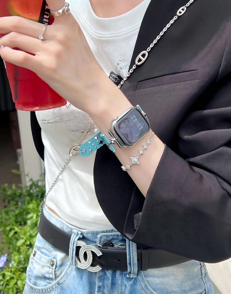 May include: A silver smartwatch with a digital display is worn on the wrist, paired with a star-shaped bracelet. A turquoise watch band and a silver chain are also visible. The person is wearing a black blazer and a black belt with a silver buckle.