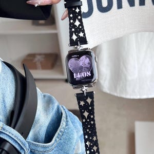 May include: A silver smartwatch with a black band featuring a star-shaped cutout design. The watch face displays the time as 14:08. The band is held by a hand with long, manicured nails.