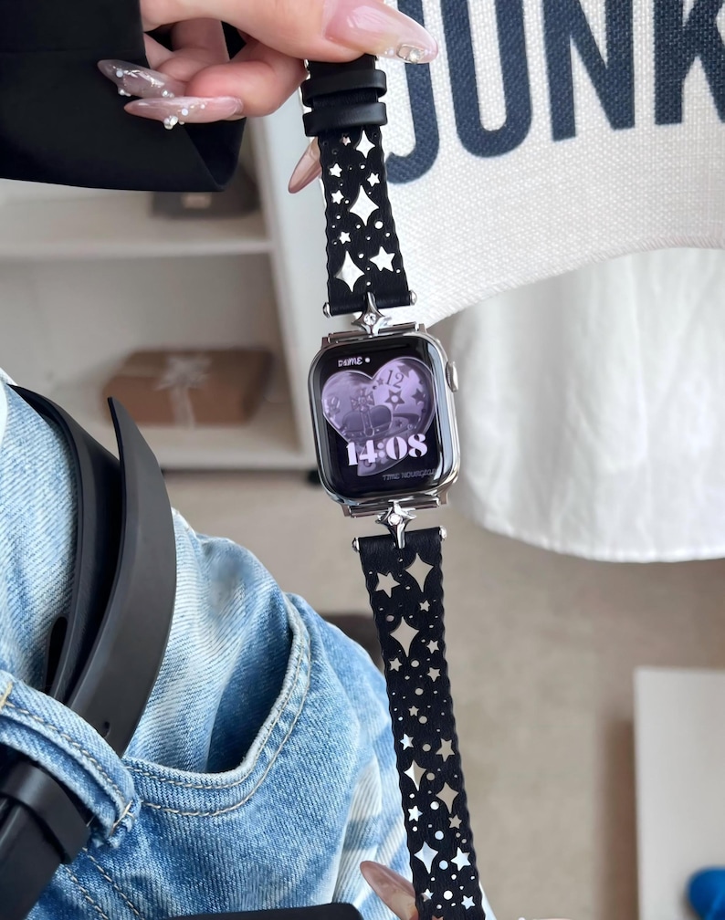 May include: A silver smartwatch with a black band featuring a star-shaped cutout design. The watch face displays the time as 14:08. The band is held by a hand with long, manicured nails.