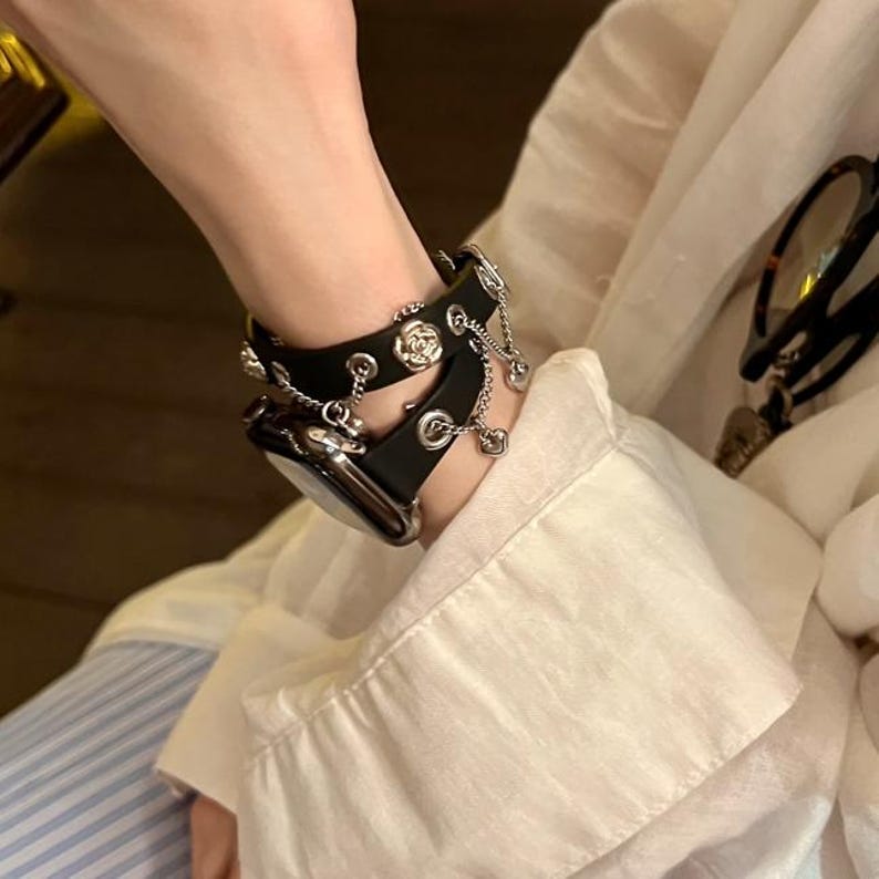 May include: Black leather watch band with silver-tone chain and floral accents. The band is designed with multiple layers and decorative elements, creating a stylish accessory. The watch band is designed to fit a smartwatch.