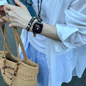 May include: A tan crocheted tote bag with a beaded necklace, a denim wallet, and a black smartwatch with a brown leather band. A black bracelet with silver accents is also visible. The person is wearing a white shirt and blue striped pants.