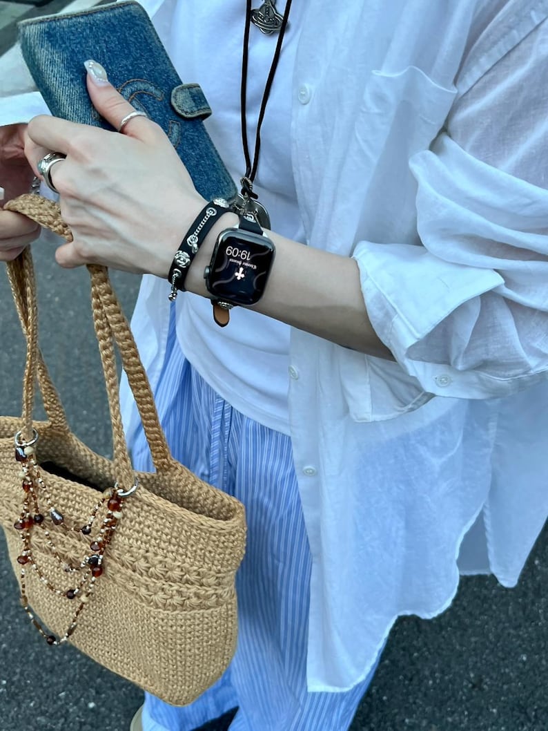 May include: A tan crocheted tote bag with a beaded necklace, a denim wallet, and a black smartwatch with a brown leather band. A black bracelet with silver accents is also visible. The person is wearing a white shirt and blue striped pants.