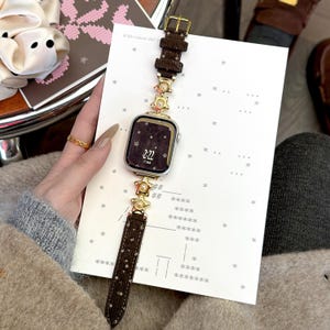 Op de afbeelding: Een smartwatch met een bruin leren bandje en goudkleurige stervormige versieringen. De wijzerplaat toont de tijd 2:22. Het horloge ligt op wit papier met sterpatronen. De band heeft een gouden gesp.