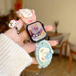 May include: An Apple Watch with a black case and a Hello Kitty display is held in a hand. The watch is surrounded by three handmade crochet accessories: a pink cat, a blue penguin, and a small white and black figurine.