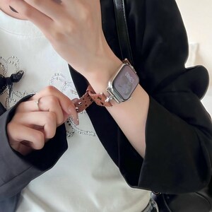 May include: Silver smartwatch with a brown patterned band. The watch face displays the time. The band features a repeating pattern of small, dark brown shapes. The watch is worn on a wrist, with a black blazer and white shirt in the background.