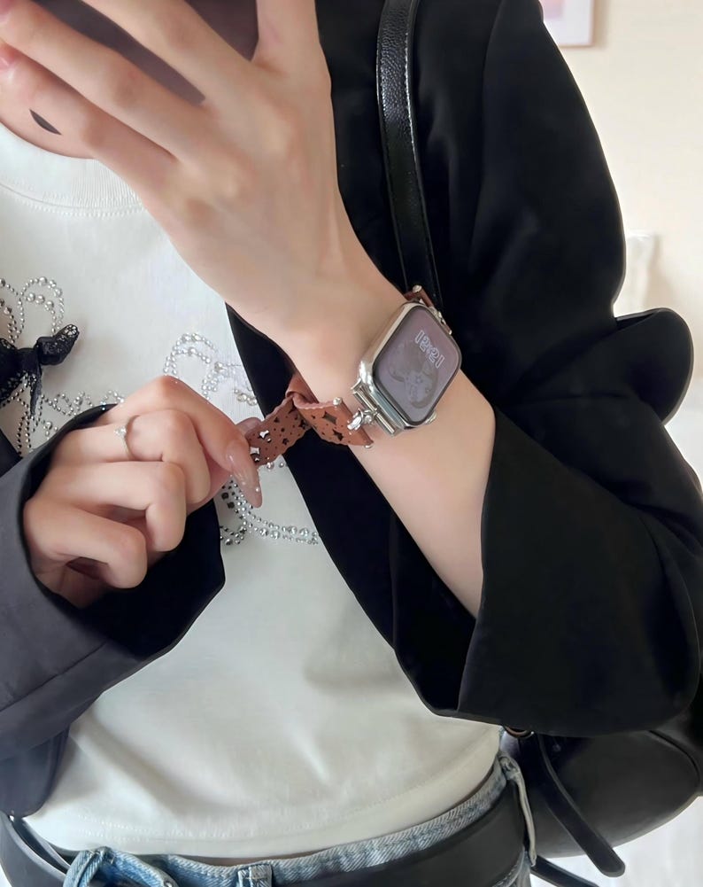 May include: Silver smartwatch with a brown patterned band. The watch face displays the time. The band features a repeating pattern of small, dark brown shapes. The watch is worn on a wrist, with a black blazer and white shirt in the background.