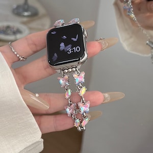 May include: A silver smartwatch with a black screen displaying the time 3:10. The watch band is adorned with iridescent butterfly charms and crystals. The watch is held by a person.