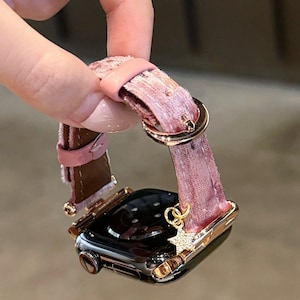 May include: A rose gold Apple Watch with a pink and brown patterned band. The band has a pink leather strap and a gold buckle. A small gold charm in the shape of a star hangs from the watch.