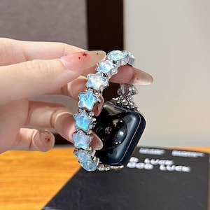 Puede incluir: Correa de reloj plateada con abalorios en forma de estrella iridiscente. La correa está sujeta a un reloj inteligente negro. Las estrellas tienen una combinación de colores azul y blanco. Un accesorio de reloj elegante.