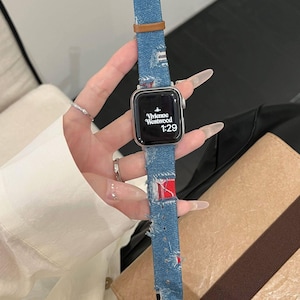 以下が含まれることがあります： シルバーのスマートウォッチ。ブルーデニムのバンドにはダメージ加工が施され、ブラウンレザーのアクセントが施されています。文字盤にはVivienne Westwoodのロゴと時刻1:29が表示されています。