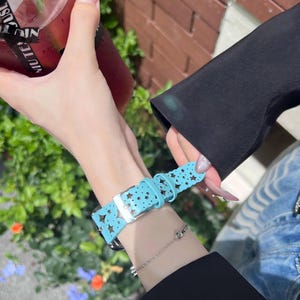May include: A light blue watch band with a star pattern. The band is made of a soft material and has a silver buckle. The watch band is worn on a wrist with a silver bracelet.