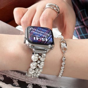 May include: A silver smartwatch with a pearl and rhinestone band is displayed. The watch face shows the time as 15:41 and the text "Vivienne Westwood CATWALK". A separate pearl bracelet is also visible.