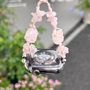May include: A silver smartwatch with a pink crystal and bead strap. The strap features large, raw rose quartz stones and smaller pink beads. The watch is suspended by a pink cord, with pink flowers in the background.