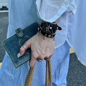 May include: A black smartwatch with a decorative leather band and silver charms. The watch displays the time as 19:11. A denim wallet and a woven tote bag are also visible.