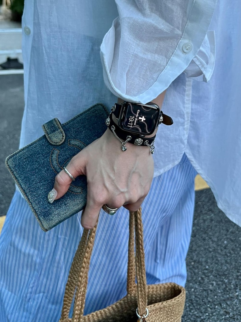 May include: A black smartwatch with a decorative leather band and silver charms. The watch displays the time as 19:11. A denim wallet and a woven tote bag are also visible.