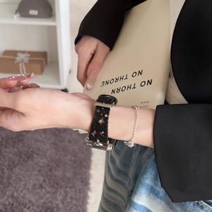 May include: Black leather watch band with a cut-out floral design. The watch band is attached to a silver watch face. A silver bracelet with a small charm is also visible.
