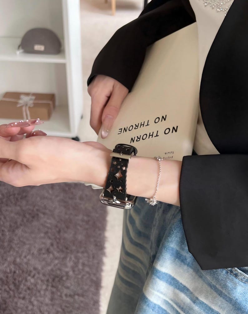 May include: Black leather watch band with a cut-out floral design. The watch band is attached to a silver watch face. A silver bracelet with a small charm is also visible.