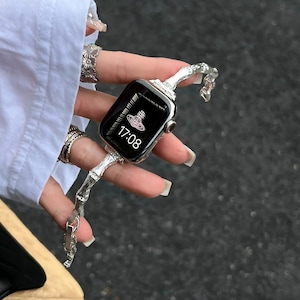 May include: Silver-toned smartwatch with a decorative band. The watch face displays the time, 17:08, and a logo. The band has a unique, textured design. The watch is held against a white shirt.