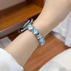 May include: A smartwatch band featuring a series of light blue, star-shaped beads with a marbled, iridescent finish. The star beads are connected by small, polished silver-toned metal spheres, creating a flexible link-style bracelet design.