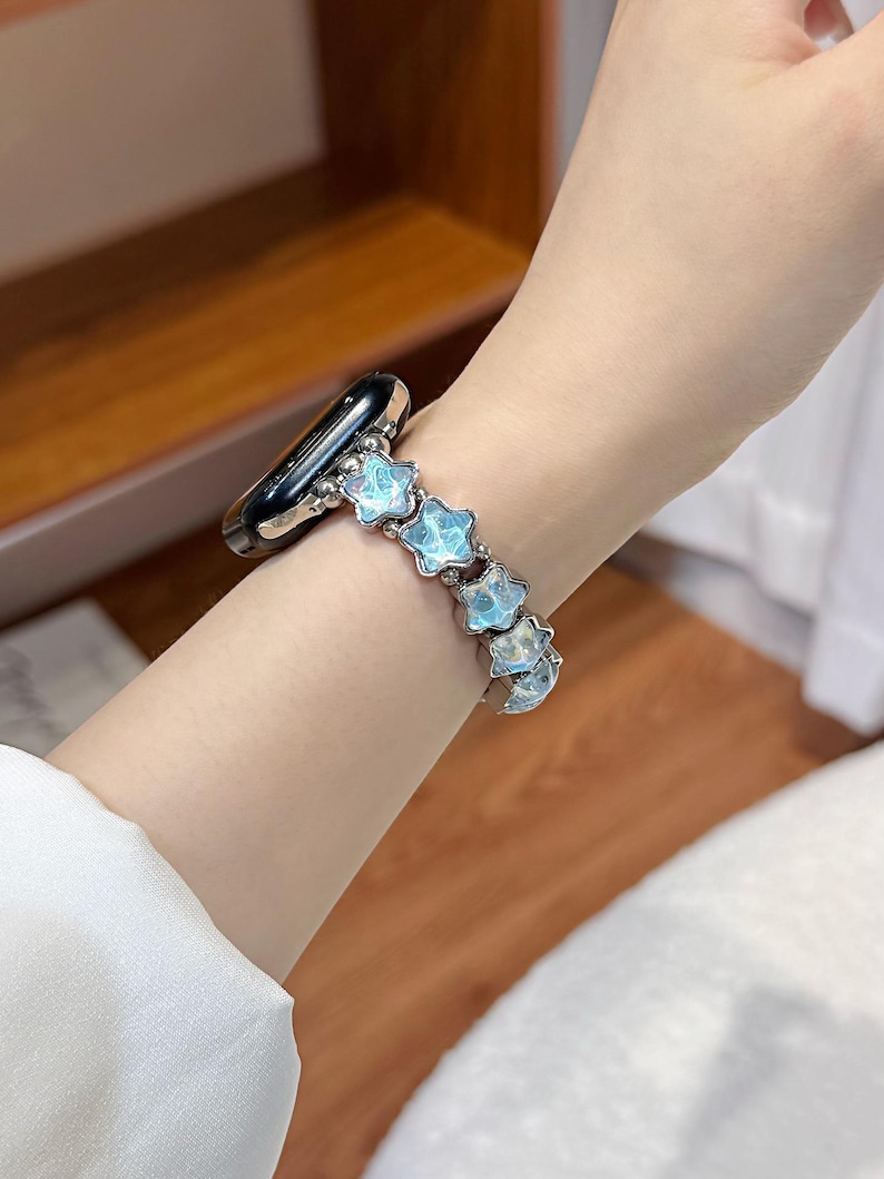 May include: A smartwatch band featuring a series of light blue, star-shaped beads with a marbled, iridescent finish. The star beads are connected by small, polished silver-toned metal spheres, creating a flexible link-style bracelet design.