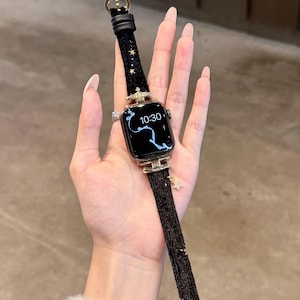 Puede incluir: Correa de reloj Apple negra con detalles dorados, incluyendo un bisel en forma de corona y dijes de estrella. La correa está hecha de un material negro texturizado y tiene una hebilla dorada.