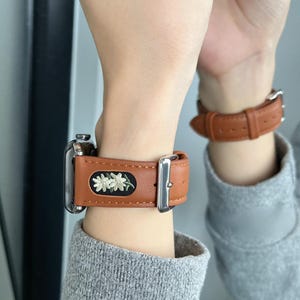 Könnte beinhalten: Braunes Leder-Uhrenarmband mit silberner Schnalle und einem schwarzen, bestickten Aufnäher mit weißen Blumen. Das Uhrenarmband ist an einem silbernen Uhrengehäuse befestigt. Ein zweites braunes Lederarmband ist sichtbar.