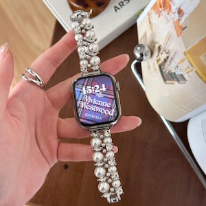 May include: A silver smartwatch with a pearl and rhinestone band. The watch face displays the time 15:24 and the Vivienne Westwood logo. The watch band is adorned with large, round pearls and sparkling rhinestones.