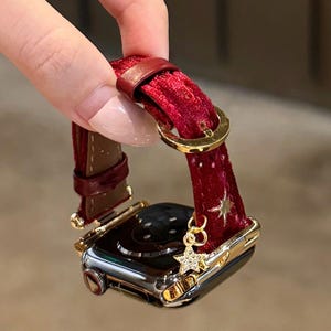 May include: A smartwatch with a burgundy velvet band and gold accents. The band features a star pattern and a dangling star charm. The watch case is silver and gold.