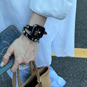 May include: A black smartwatch with a leather band and decorative silver charms is worn on a wrist. The watch face displays the time as 19:10. A denim wallet and a woven handbag with beaded accents are also visible.