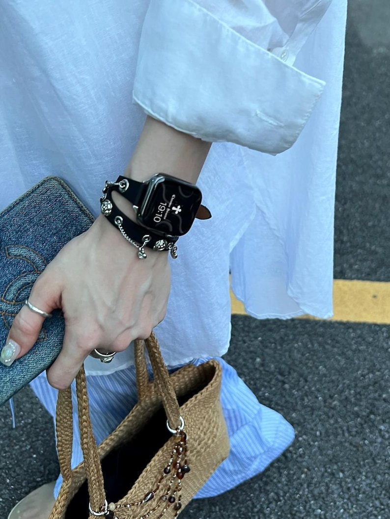 May include: A black smartwatch with a leather band and decorative silver charms is worn on a wrist. The watch face displays the time as 19:10. A denim wallet and a woven handbag with beaded accents are also visible.