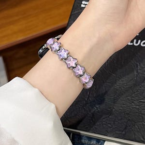 May include: A watch band featuring a series of linked, star-shaped charms with a pearlescent pink and purple finish. The star charms are connected by small silver-toned metal beads, creating a flexible bracelet design that attaches to a watch face.